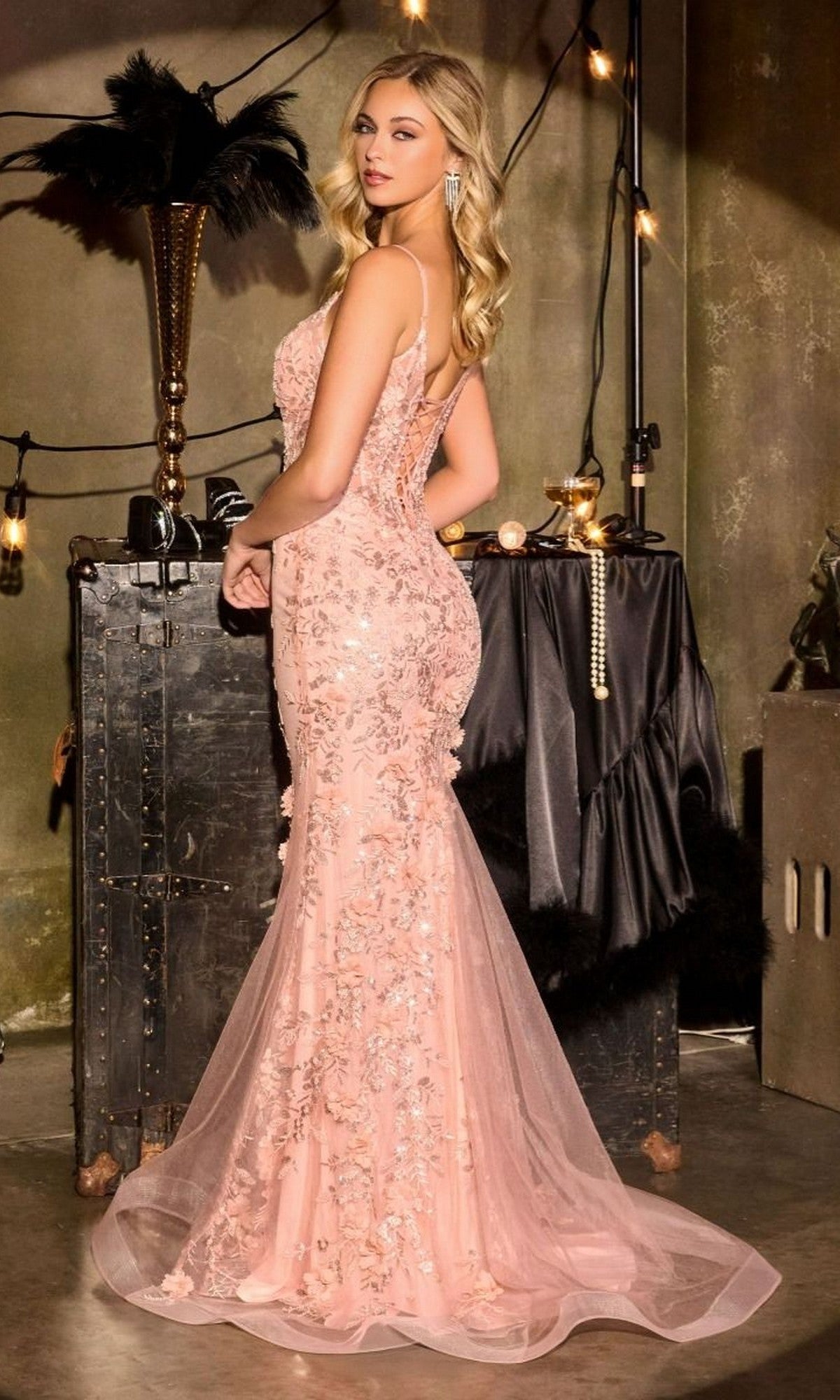 A woman stands indoors facing slightly away, wearing the Ladivine CT264— a fitted, blush pink mermaid prom dress with sequins, sheer corset bodice, and vintage-style decor. String lights twinkle softly in the background.